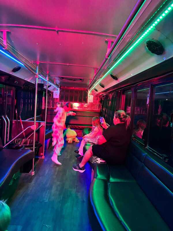 Passengers dance and relax inside a bus lit with vivid pink and green LEDs, creating a lively, playful party atmosphere.