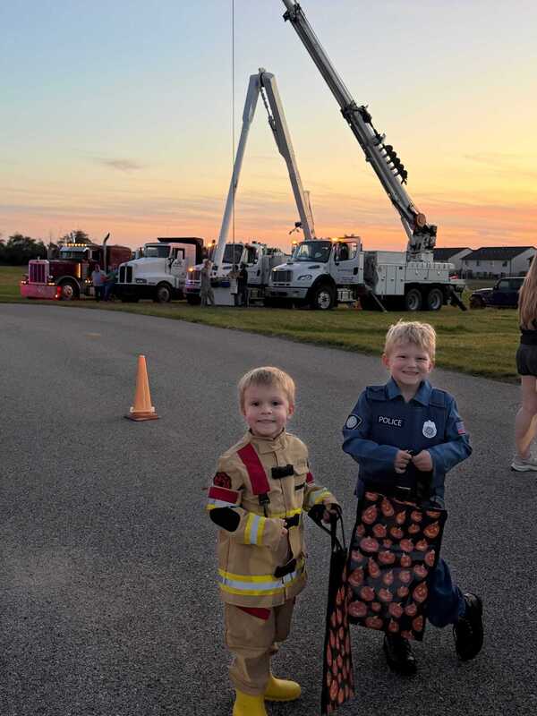 Two young children in firefighter and police costumes pose beside a road, with fire trucks and a towering crane display behind them at sunset.