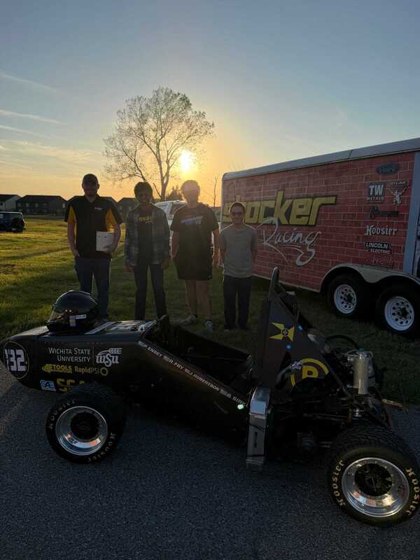 Four people stand behind a student-built race car at sunset, with a branded trailer in the background; warm backlighting creates a proud, celebratory mood.