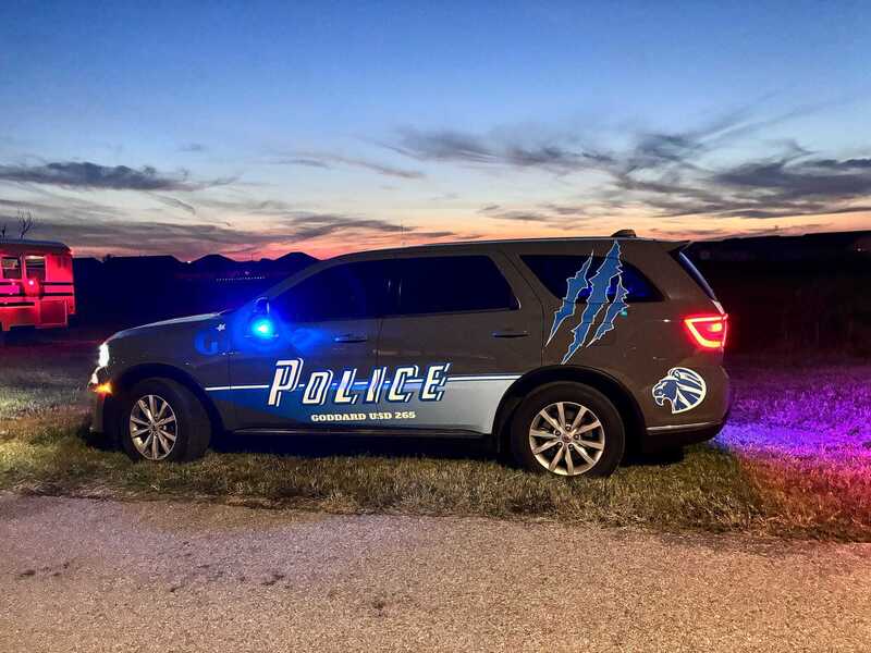 A marked police SUV with blue emergency lights and a mountain-themed decal is parked beside a rural road at sunset, casting blue and red light.