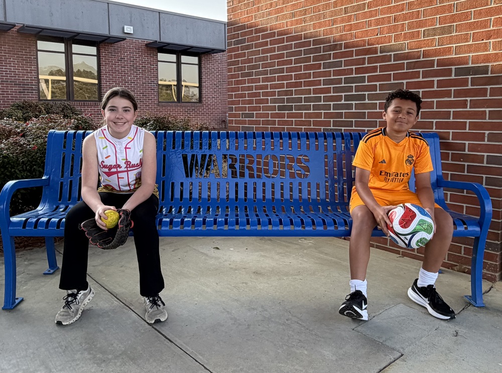 Two students sit on a blue “Warriors” bench; one holds a softball and glove, the other holds a soccer ball.