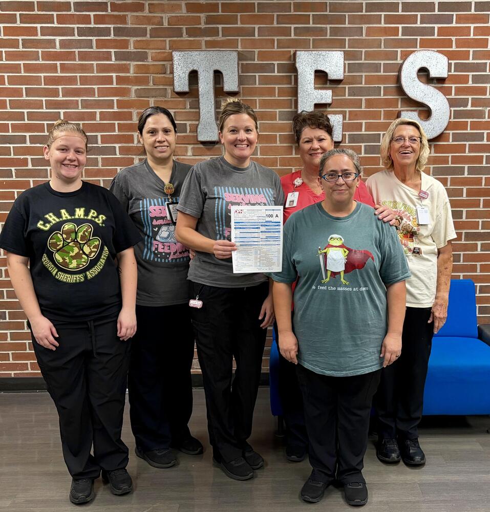 Six Tolbert Elementary nutrition staff members pose together in front of a brick wall with large “TES” letters. One staff member holds a health inspection report showing a perfect score of 100/A. The group smiles proudly while celebrating the achievement.