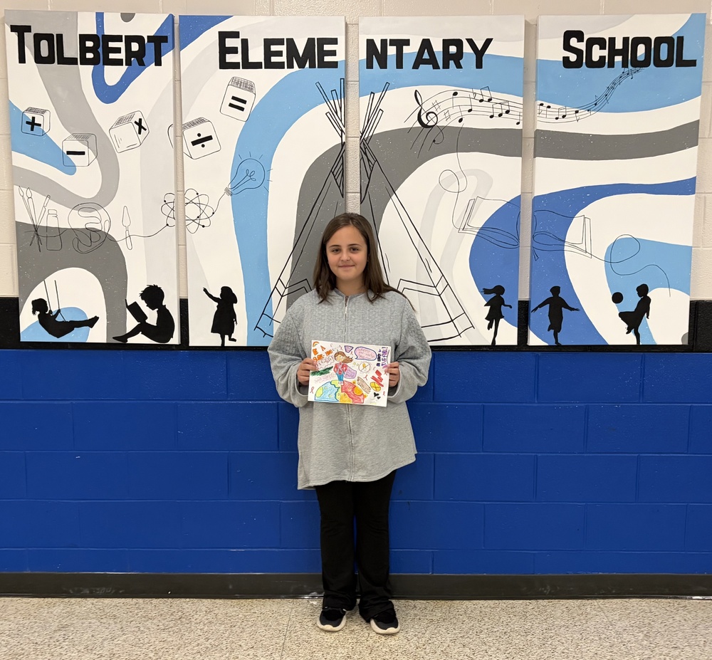 A student smiles while holding a superhero-themed drug prevention poster in front of the Tolbert Elementary School mural.