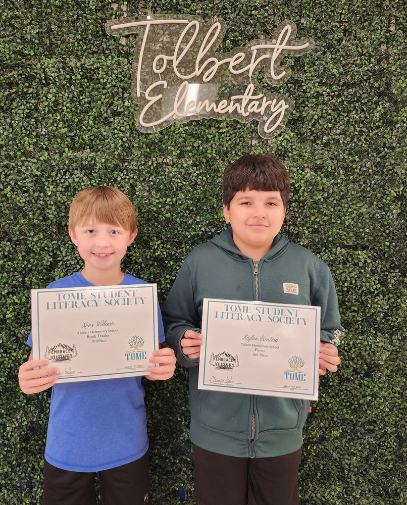Two elementary school boys stand in front of a green leafy backdrop with a sign reading “Tolbert Elementary.” Each is smiling and holding a certificate from the TOME Student Literacy Society. The boy on the left wears a blue shirt and holds an award for a book trailer, while the boy on the right wears a green hoodie and holds an award for a poem.