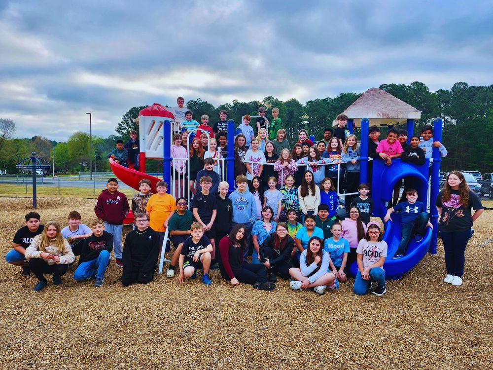 A large group of elementary or middle school students pose together on a colorful playground structure outdoors. The children are arranged in rows—some standing on the play equipment, others sitting or kneeling in front—smiling toward the camera. The playground features red and blue slides and climbing structures, with wood chips covering the ground. In the background, there are trees, a cloudy sky, and parts of a park with fences and equipment visible.