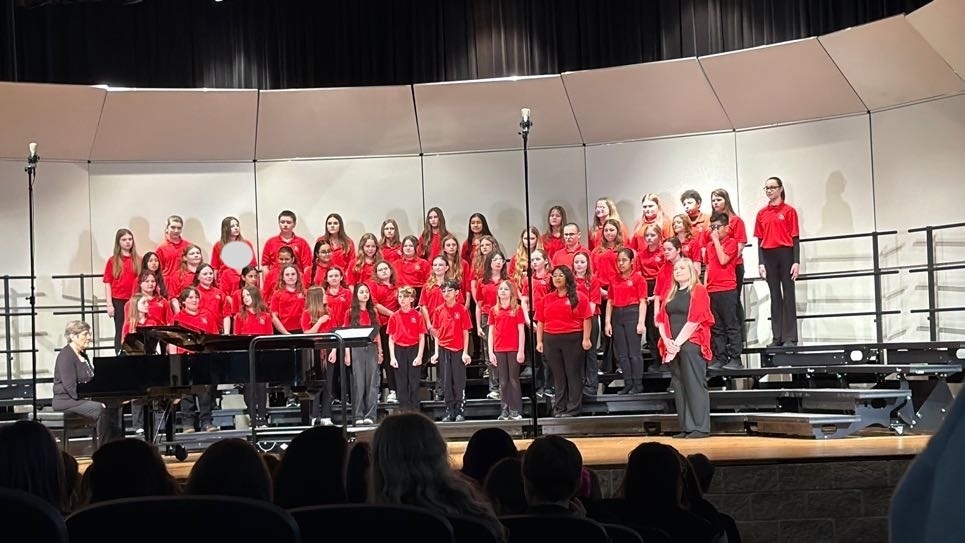 A group of students standingi on risers at a choral concert. They are wearing red polo shirts with black pants.