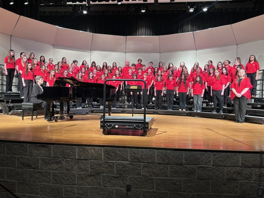 A group of students wearing red tops and black pants are singing on risers.