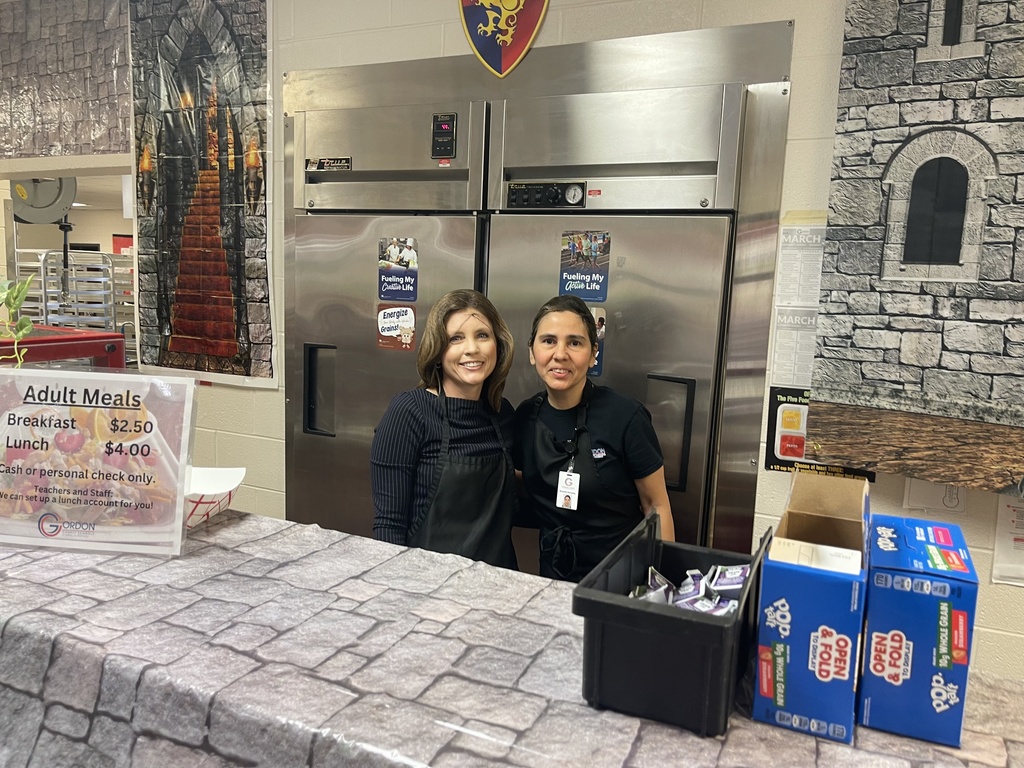 County Superintendent Alice Mashburn stands behind the cafeteria serving counter with a school nutrition staff member. The space is decorated with medieval castle backdrops, shields, and stone wall coverings for a themed breakfast event.