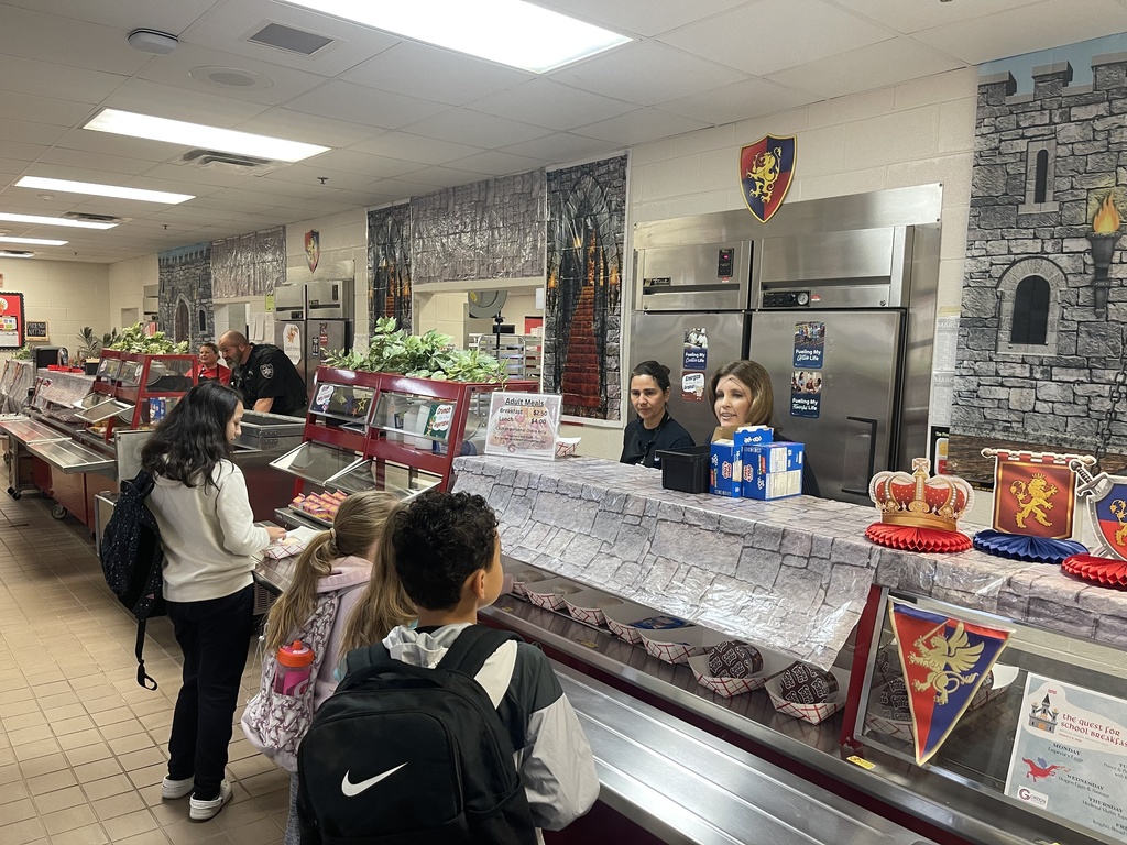 Students with backpacks move through a medieval-themed breakfast line in the school cafeteria. Superintendent Alice Mashburn and cafeteria staff serve food behind a counter decorated with stone-patterned coverings, crowns, and royal banners. A sheriff’s deputy assists further down the line.