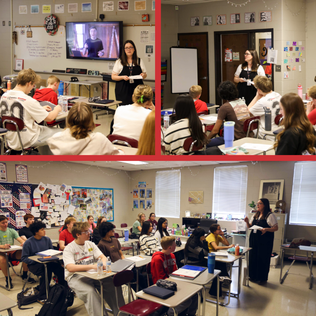 A teacher stands at the front of a high school classroom speaking to students seated at desks. A video is displayed on a screen behind her while students take notes and listen attentively. The classroom walls are decorated with posters and student work.