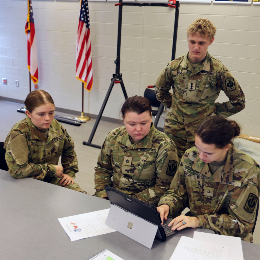 Alt Text: Four JROTC students in camouflage uniforms collaborate at a table while one student types on a laptop and others look on. Papers are spread out on the table, and American and state flags stand in the background of the classroom.