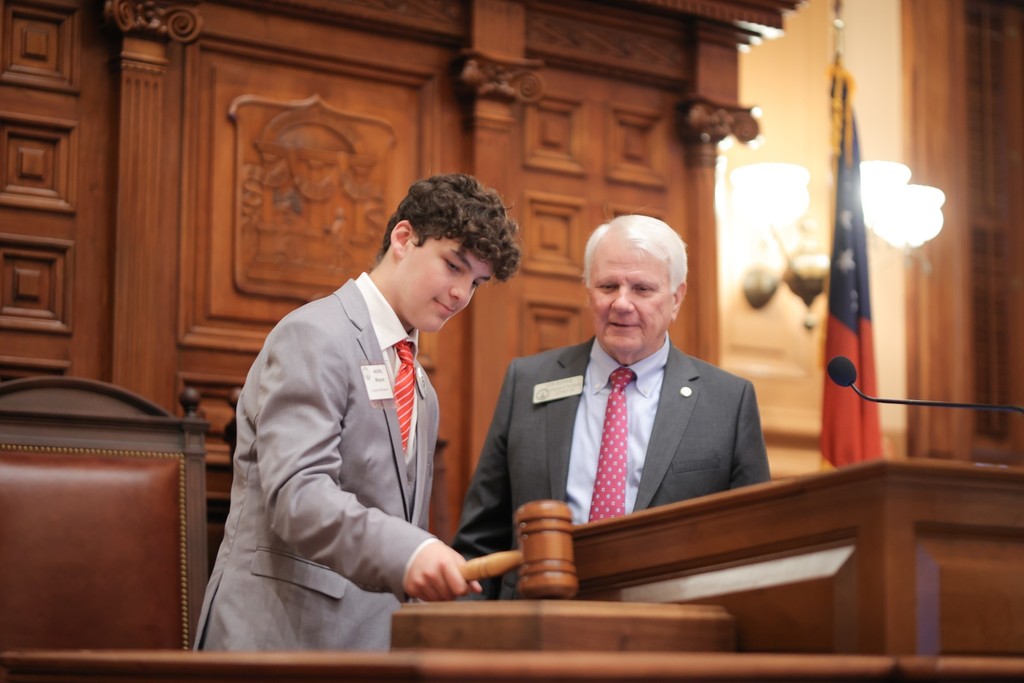 Student striking a large gavel on a desk beside a state representative at the Capitol.