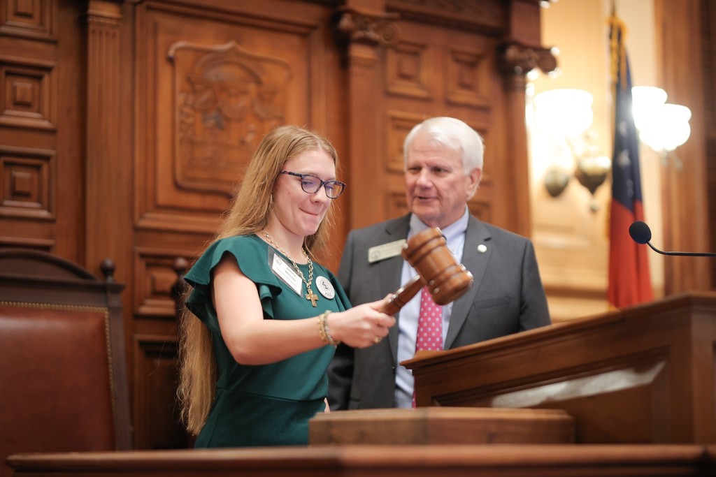 Student striking a large gavel on a desk beside a state representative at the Capitol.