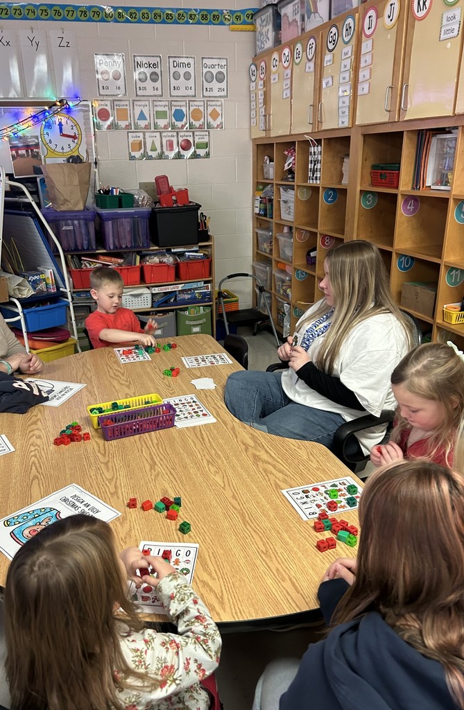 A paraprofessional works closely with a small group of students at a table, offering guidance and support as they complete an activity. The students are engaged and focused.