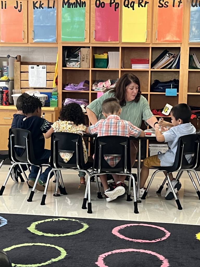 A paraprofessional works closely with a small group of students at a table, offering guidance and support as they complete an activity. The students are engaged and focused.