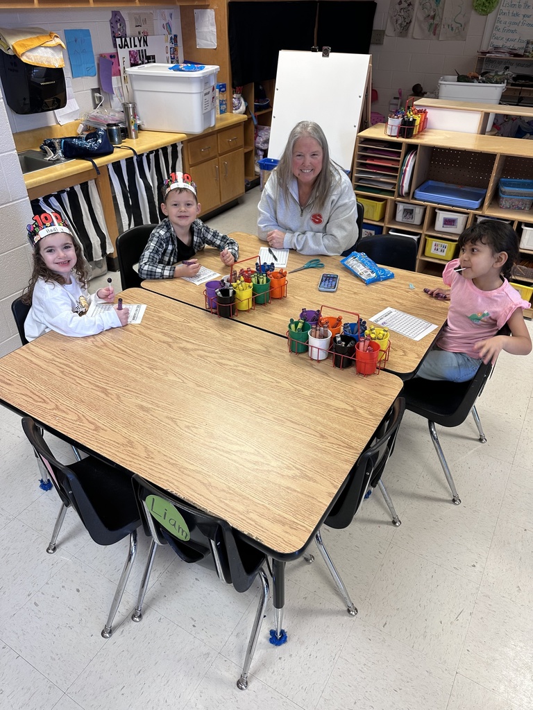 A paraprofessional smiles while assisting a student one-on-one, helping them with their work. The classroom environment is bright and welcoming.