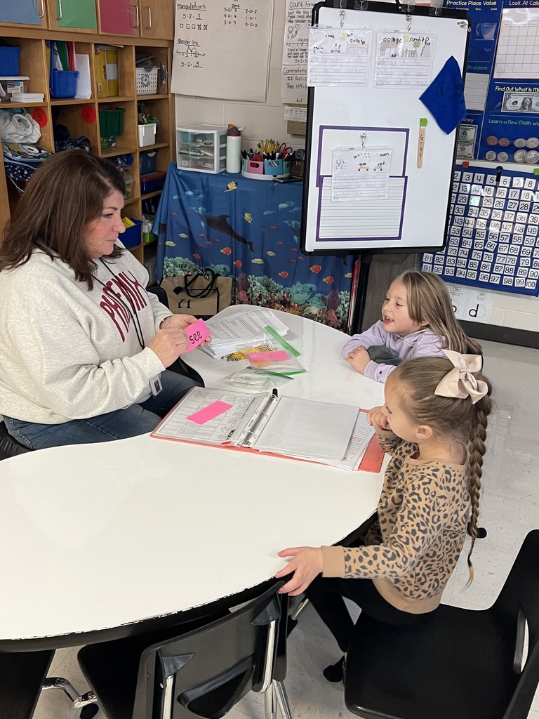 A paraprofessional helps students during a hands-on activity, with materials spread out on a table. The students appear excited and involved.