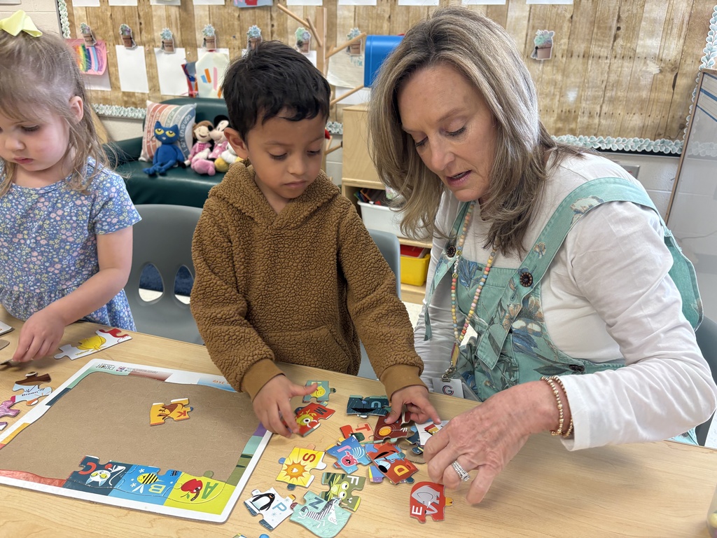 A paraprofessional helps students during a hands-on activity, with materials spread out on a table. The students appear excited and involved.