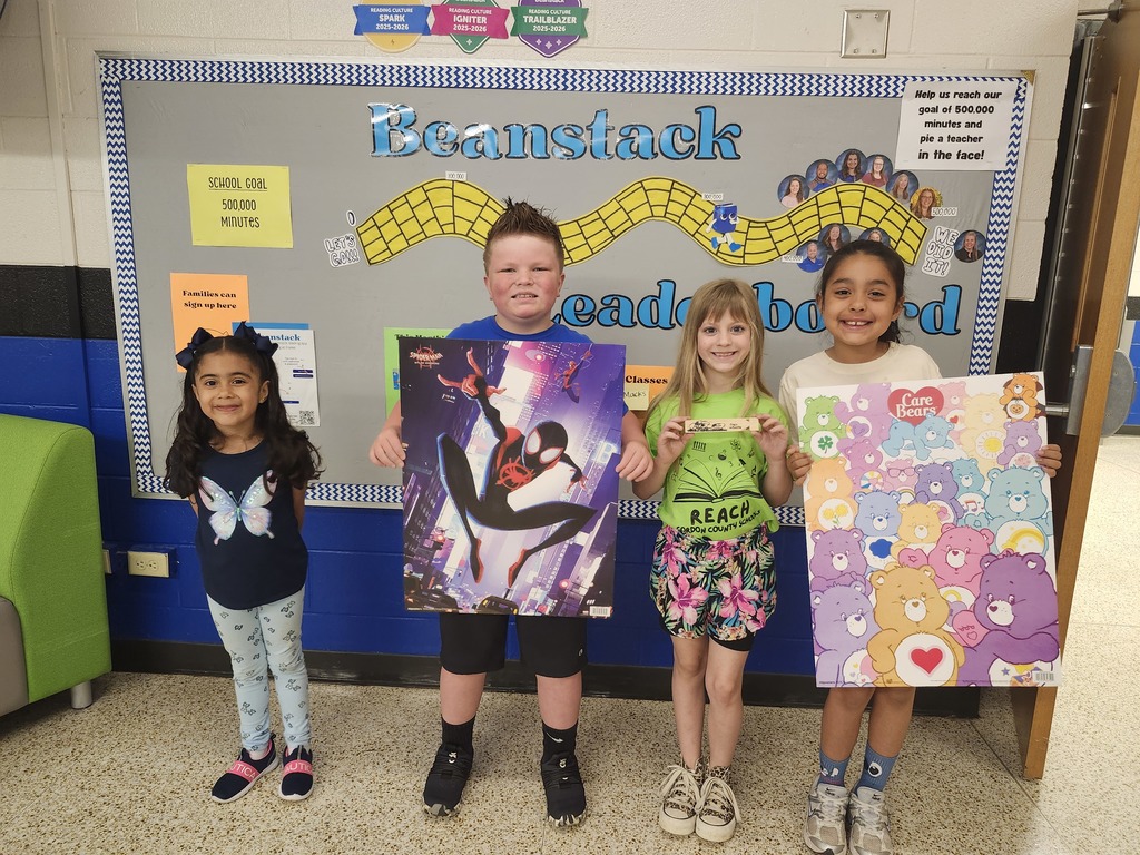 Four elementary students stand smiling in front of a “Beanstack Leaderboard” display at school. Two students hold large posters—one of Spider-Man and one of Care Bears—while the others stand proudly beside them. The bulletin board behind them highlights a school reading goal of 500,000 minutes.
