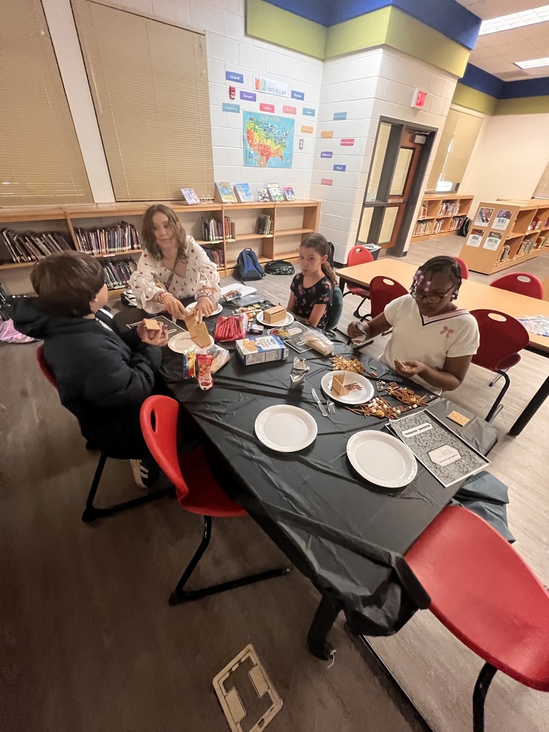 Four students sit at a table covered in black paper, carefully constructing edible houses from graham crackers and frosting. Bookshelves and a map display are visible in the background of the library.