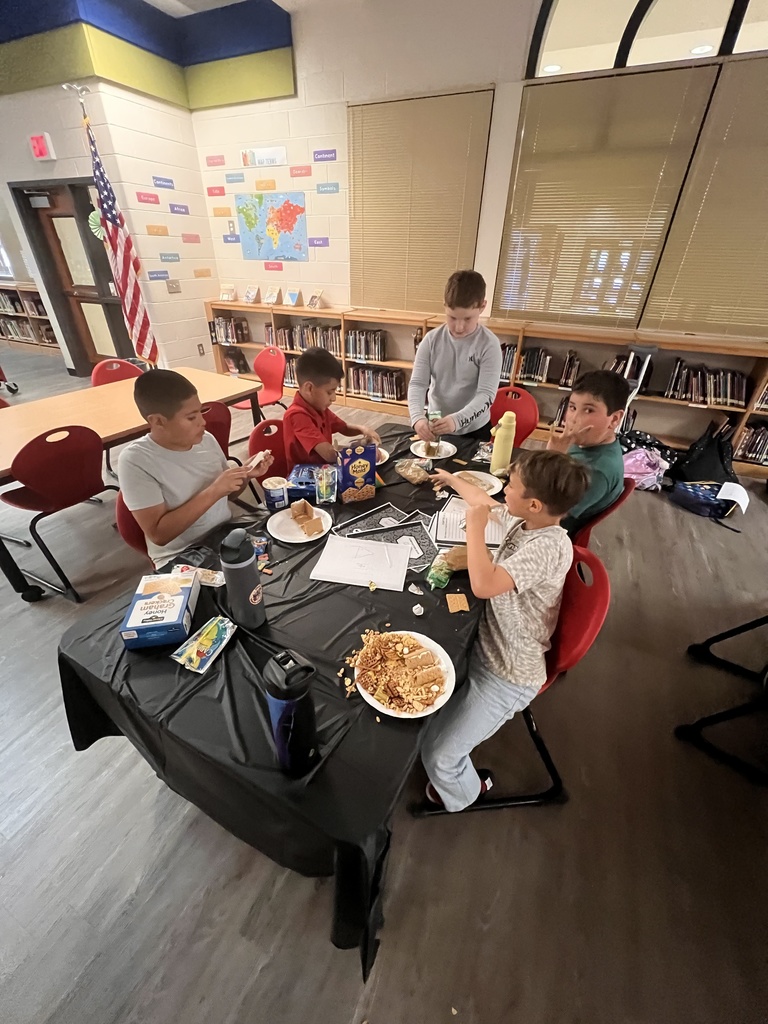 Five students sit and stand around a table in the library, working together to build edible book-themed houses. Plates of graham crackers, icing, and snacks are spread across the table as they assemble their designs.