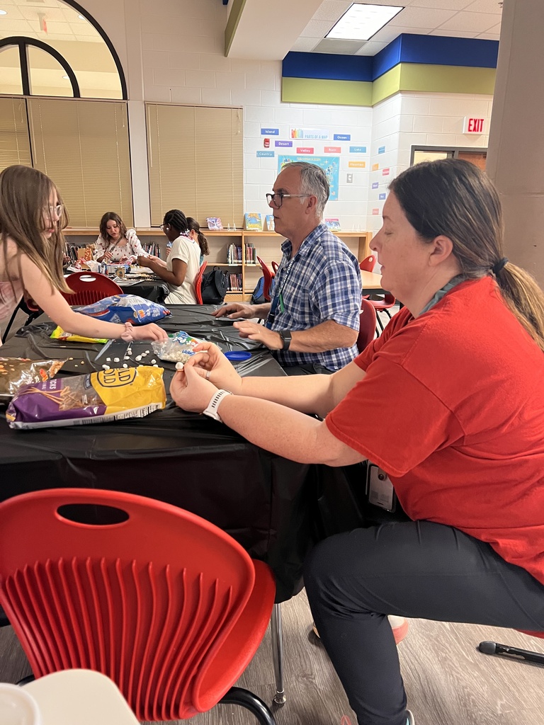 Two adults sit at a table alongside a student, helping sort and place small marshmallows and candy pieces for an edible house project. Other students work at tables in the background.