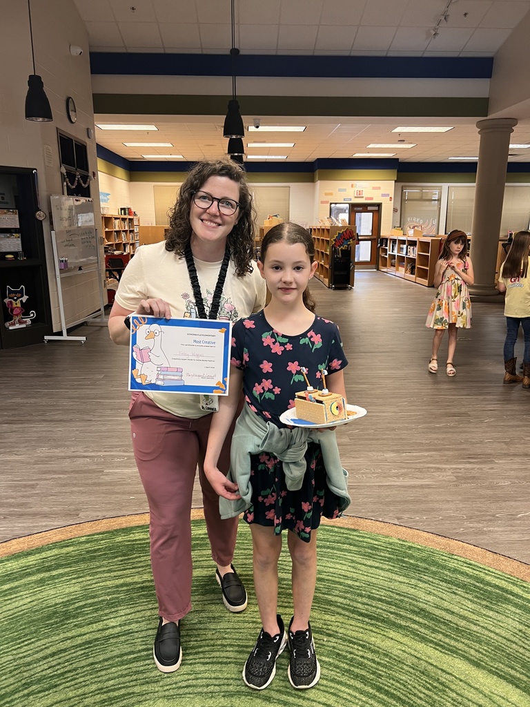 A teacher stands next to a student holding a certificate labeled “Most Creative.” The student smiles while holding their award in the library.