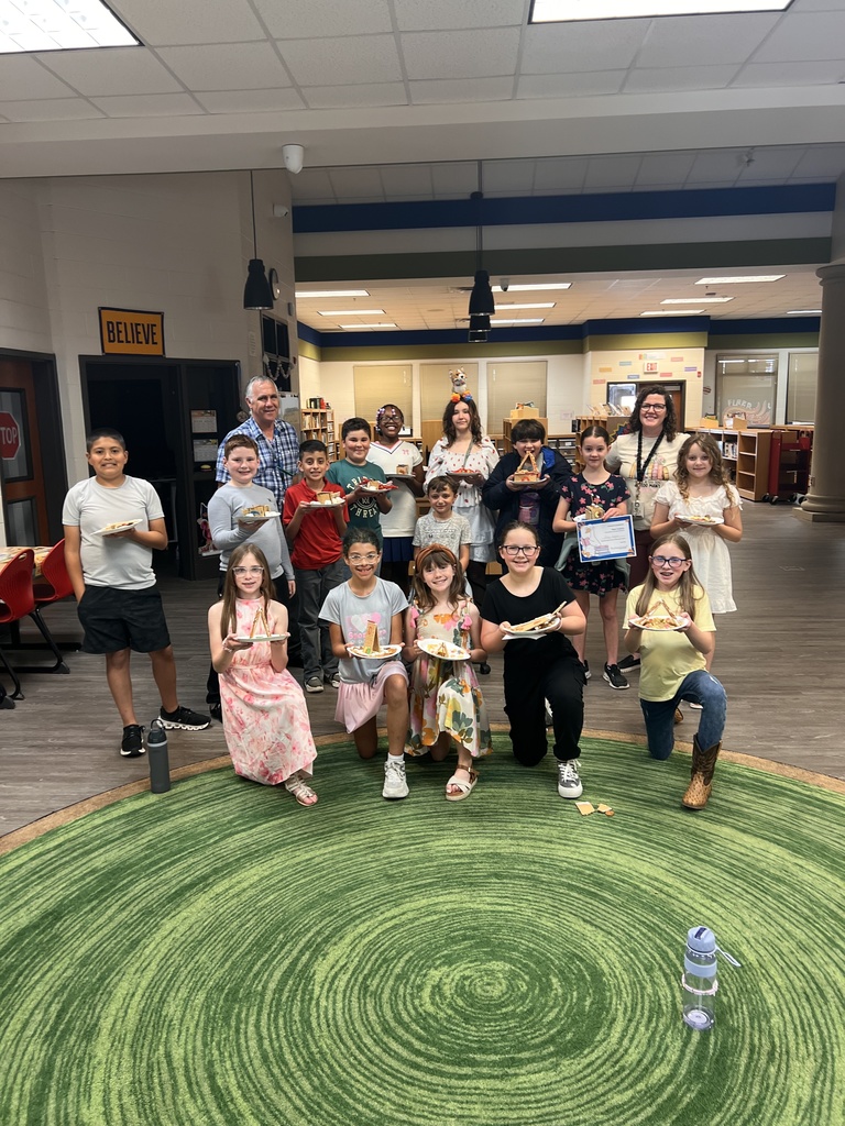 A teacher stands beside a student holding a plate with an edible house. The teacher holds a certificate labeled “Most Creative,” and both are smiling in the school library.