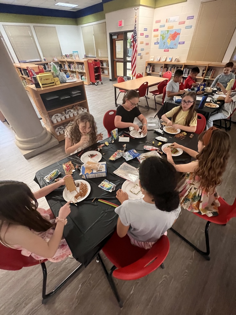 A group of elementary students sit around a table in the school library, building edible houses using graham crackers, pretzels, frosting, and candy. The table is covered with supplies, and students are focused on decorating their creations.