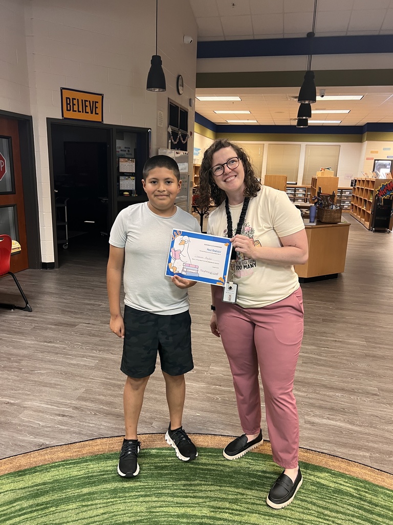 A teacher stands next to a student holding a certificate labeled “Most Realistic.” The student smiles while holding their award in the library.