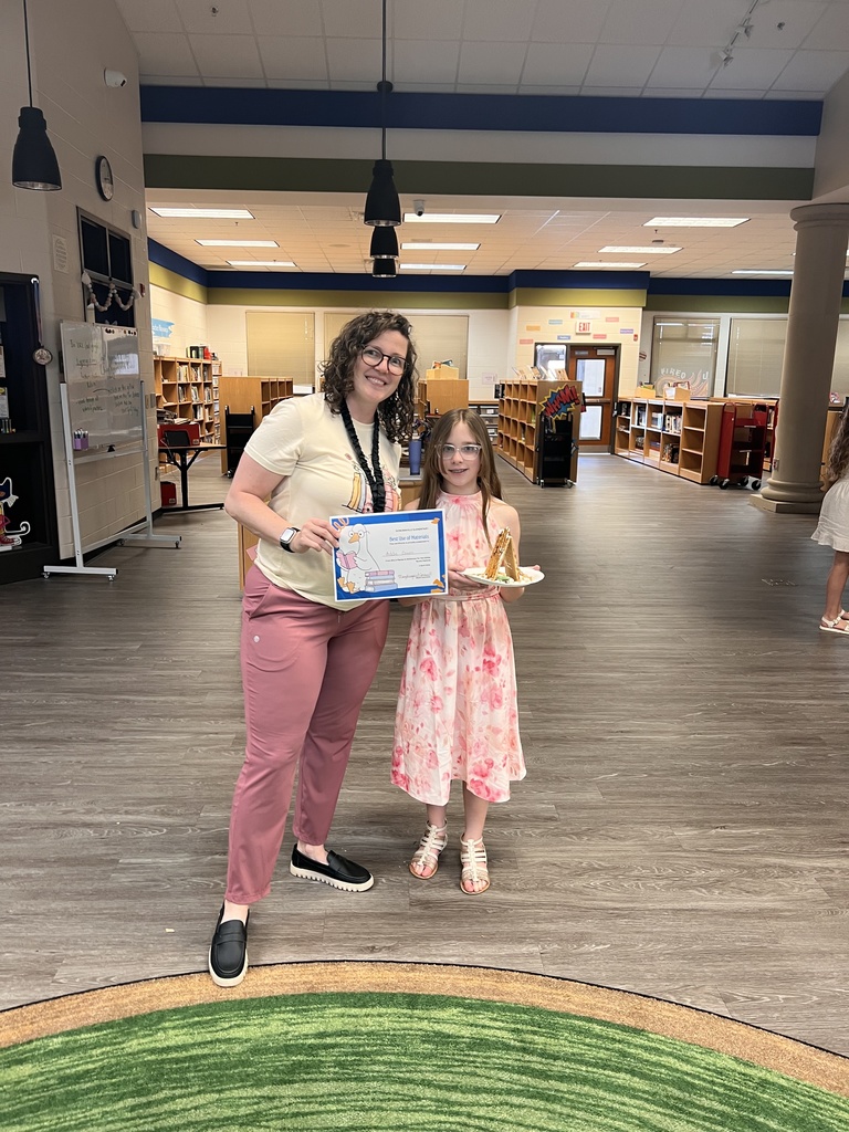 A teacher poses with a student holding a certificate labeled “Best Use of Materials.” The student holds their edible house while smiling for the photo.