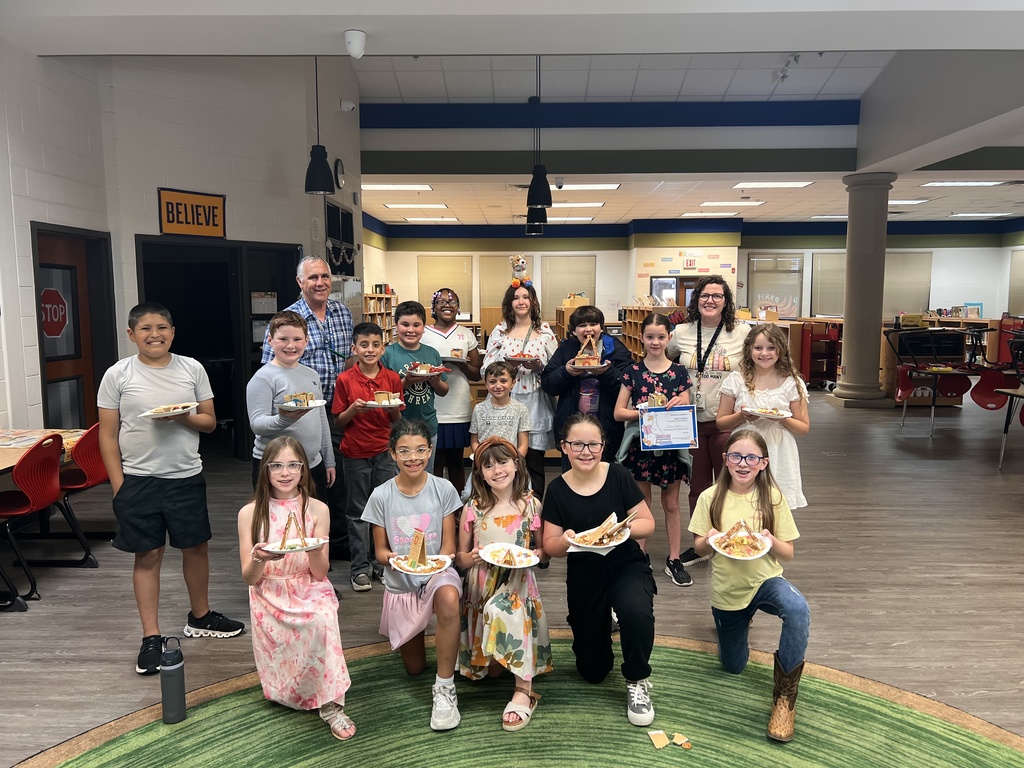 A group of students and teachers pose together in the library, smiling and holding their completed edible house creations on plates. The group stands on a green circular rug.