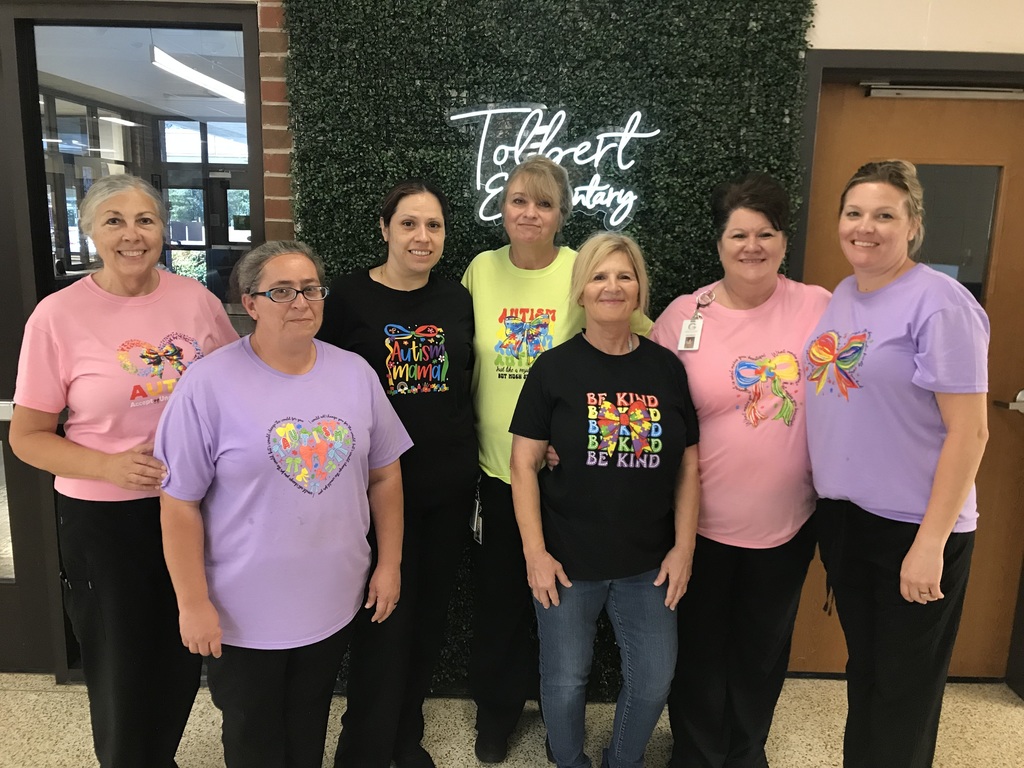A group of seven women stand side by side indoors in front of a green plant wall with a lit sign that reads “Tolbert Elementary.” They are smiling and wearing colorful T-shirts with autism awareness designs, including puzzle pieces, hearts, and phrases like “Be Kind” and “Autism Mama.” The setting appears to be a school hallway with doors and windows visible on either side