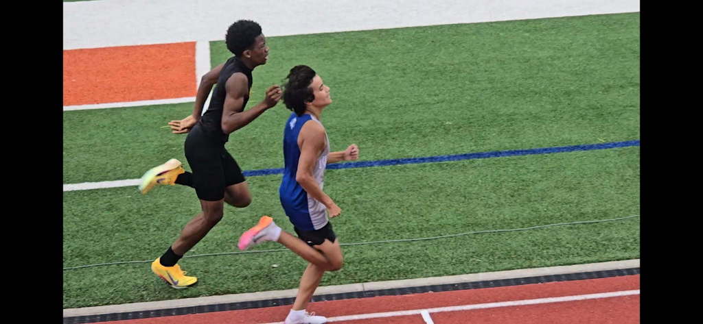 Two athletes running side by side on a track during a race, captured from above, with the inside runner slightly ahead and field turf visible beside the track.