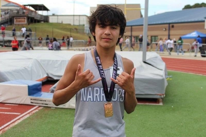 Student athlete standing on a track field wearing a Warriors uniform and a medal, holding both thumbs up in a celebratory gesture, with track equipment and spectators in the background.