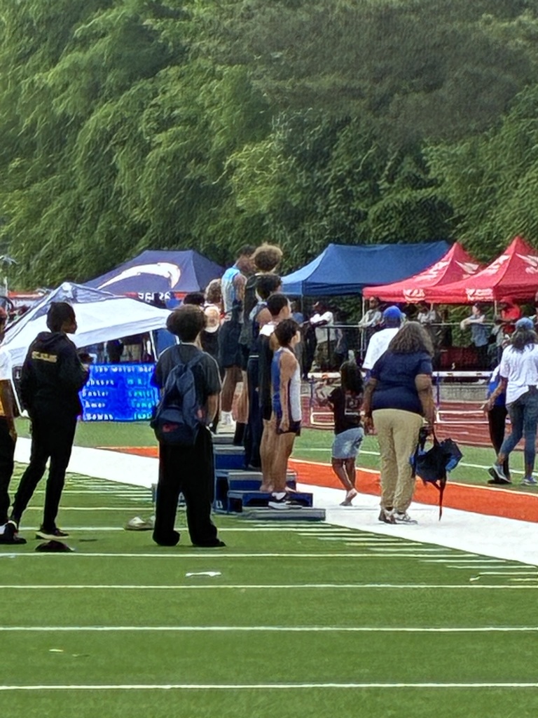 Athletes standing on a podium during an award ceremony on a track field, with event tents, spectators, and trees visible in the background.