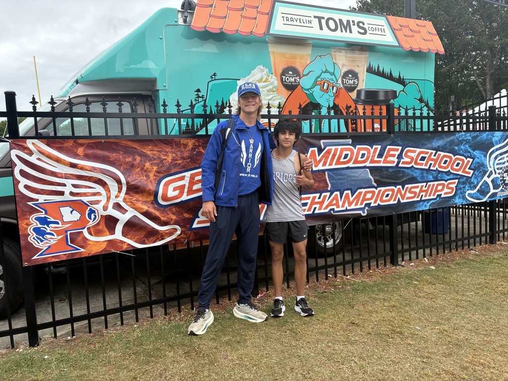 Two people standing in front of a fence with a banner reading “Middle School Championships,” posing near a colorful food truck, with grass and outdoor event surroundings visible.
