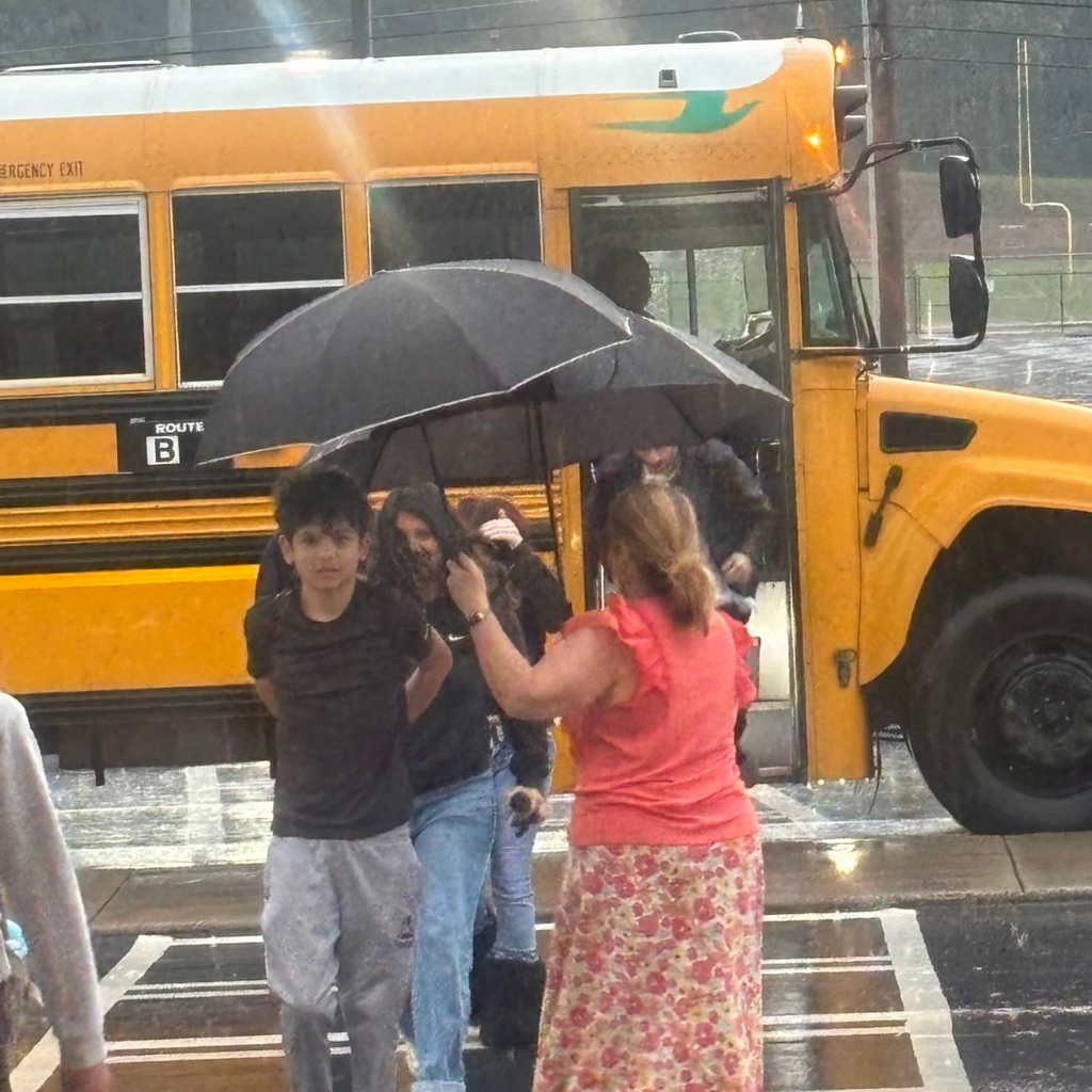 Mrs. Shannon Rainwater holding two umbrellas over students getting off the bus this morning during rainy weather.