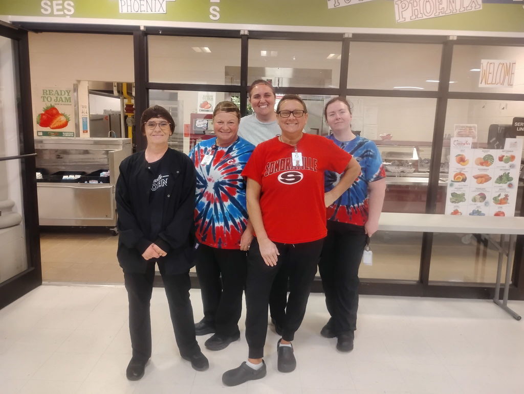Five lunchroom ladies standing in front of a folding table. They are in front of a glass windows. All are wearing red and black and have thier hair pulled back. 