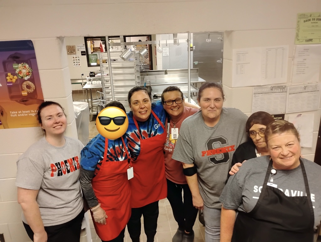 Image of seven lunchroom ladies in front of the door to the kitchen. All are wearing Sonoraville Elementary School tshirts and have their hair pulled back. 