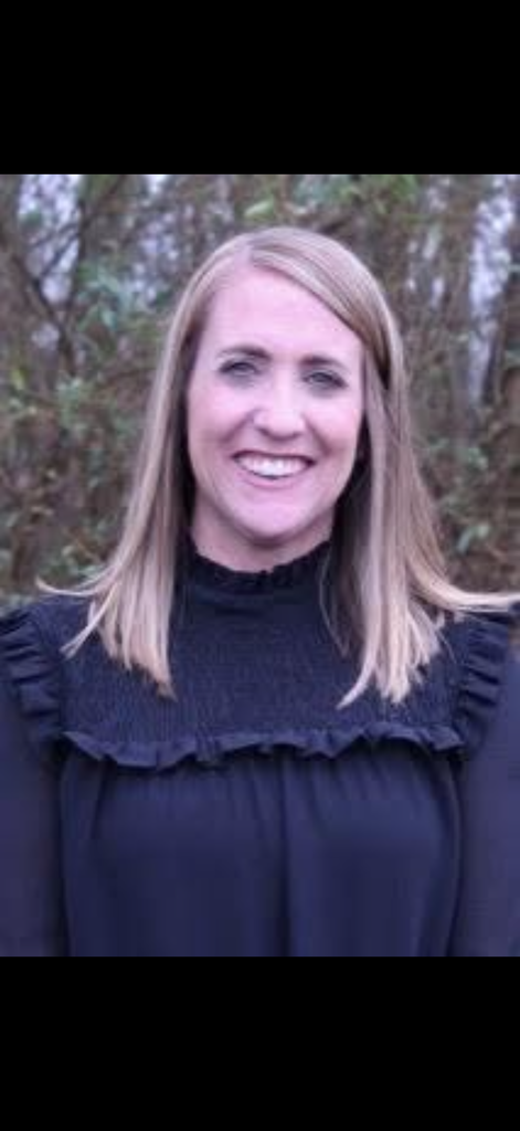 Photo of Mrs. Kelly Rampy smiling in a black top in front of a wooded area. 