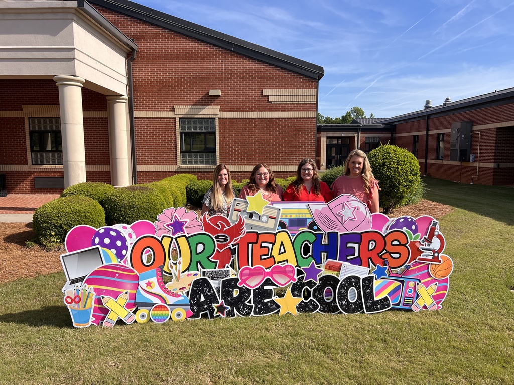 Four first grade teachers all dressed in grey tshirts stand behind a yard sign reading "our teachers are cool"