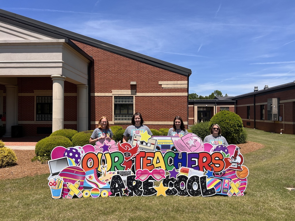 Four Fourth grade teachers all dressed in grey tshirts stand behind a yard sign reading "our teachers are cool"