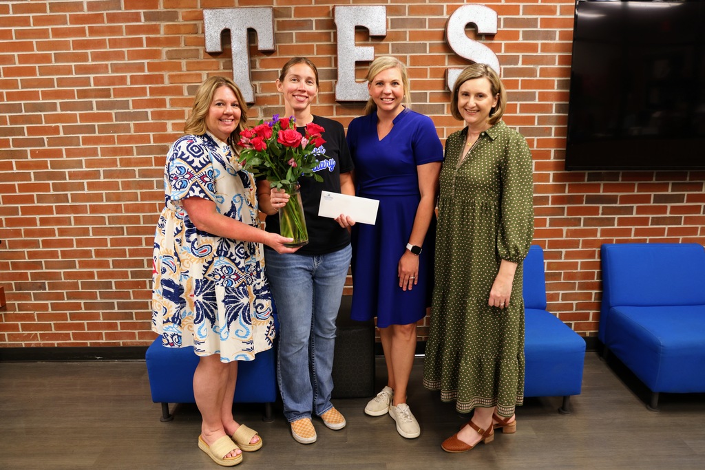Kim Gallman, Emily Rossberg, Dr. Rebecca Owen, and Mendy Goble standing together with flowers and a check to celebrate District Teacher of the Year