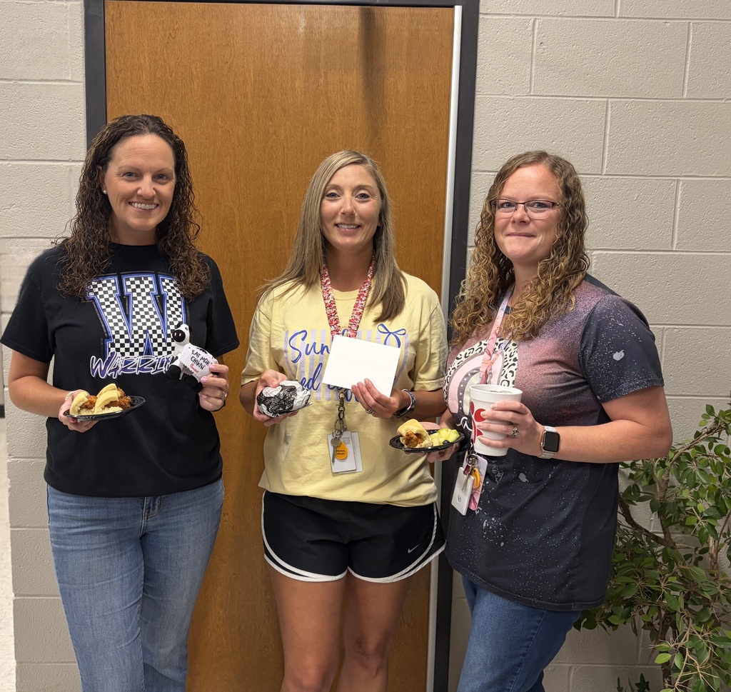 Three smiling adults stand in front of a wooden door inside a school hallway, each holding a plate of food. The person on the left wears a black school spirit T-shirt and jeans, the person in the middle wears a light yellow T-shirt and black athletic shorts while holding a wrapped sandwich and an envelope, and the person on the right wears a dark graphic T-shirt and jeans while holding a drink. All three are wearing staff ID badges, suggesting they are enjoying a staff appreciation breakfast 