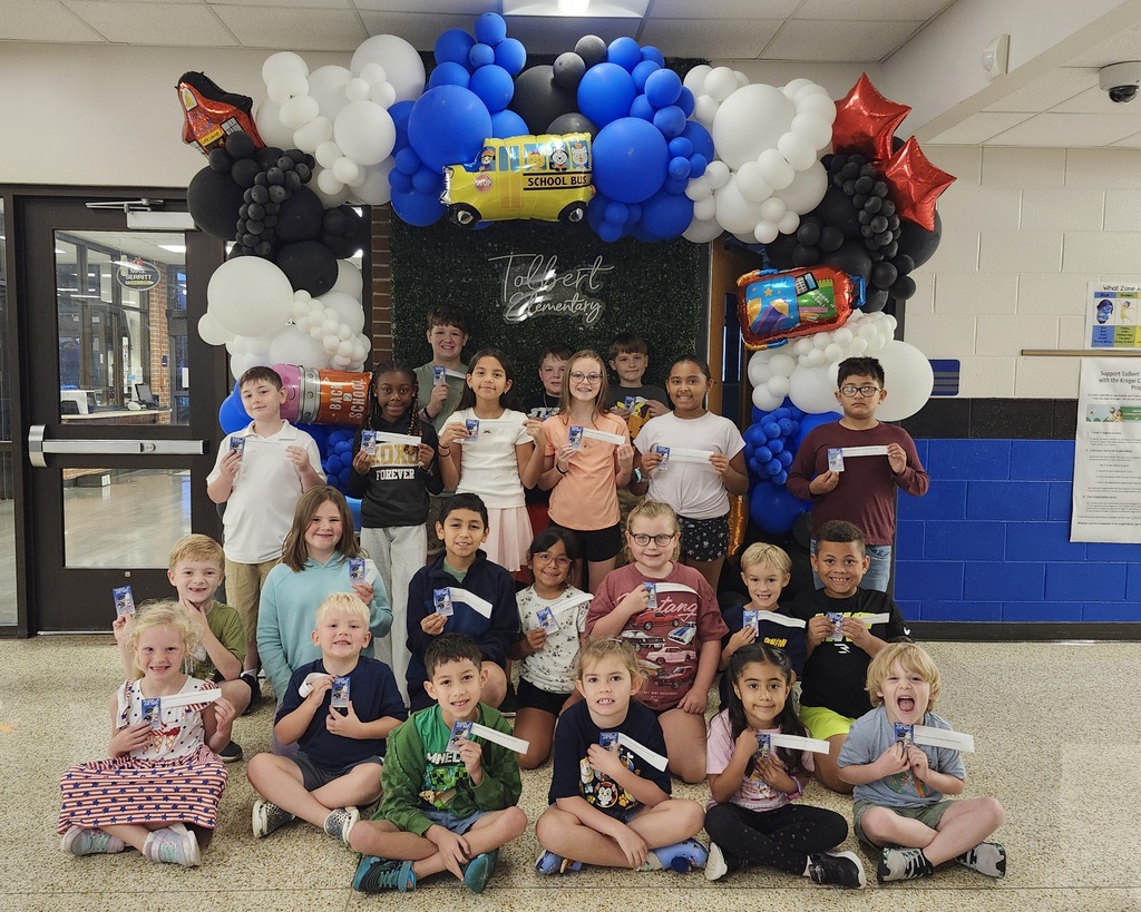 Group of elementary students posing in a school hallway while holding small cards and envelopes. Behind them is a “Tolbert Elementary” backdrop surrounded by a large balloon arch in blue, white, and black, with colorful school-themed balloons.