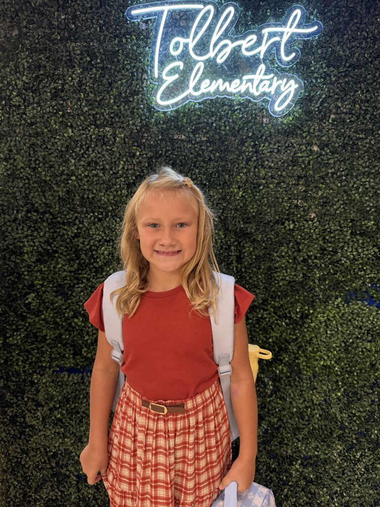 A smiling student with a backpack stands in front of a greenery wall beneath a glowing “Tolbert Elementary” sign.