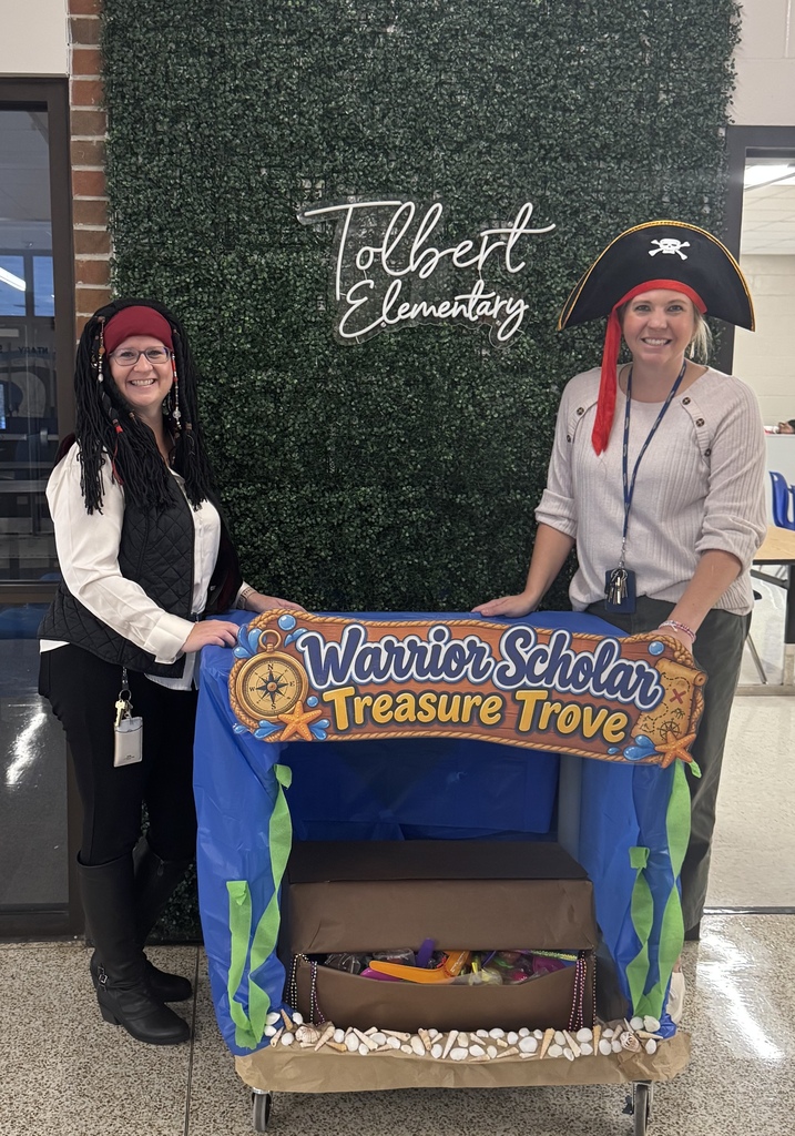Two Tolbert Elementary staff members dressed as pirates stand beside the “Warrior Scholar Treasure Trove” treat cart in front of a Tolbert Elementary greenery wall.
