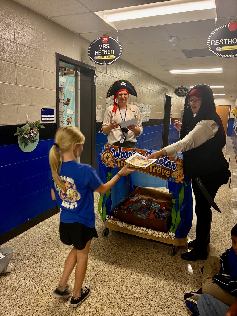Two Tolbert Elementary staff members dressed as pirates present a certificate to a student beside the Warrior Scholar Treasure Trove treat cart in the school hallway.