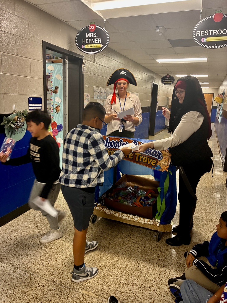Two Tolbert Elementary staff members dressed as pirates recognize students and hand out certificates beside the Warrior Scholar Treasure Trove treat cart in the school hallway.